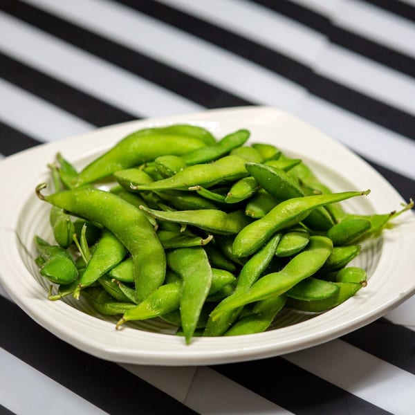 A white bowl filled with vibrant green edamame pods, lightly salted, on a striped black and white surface.