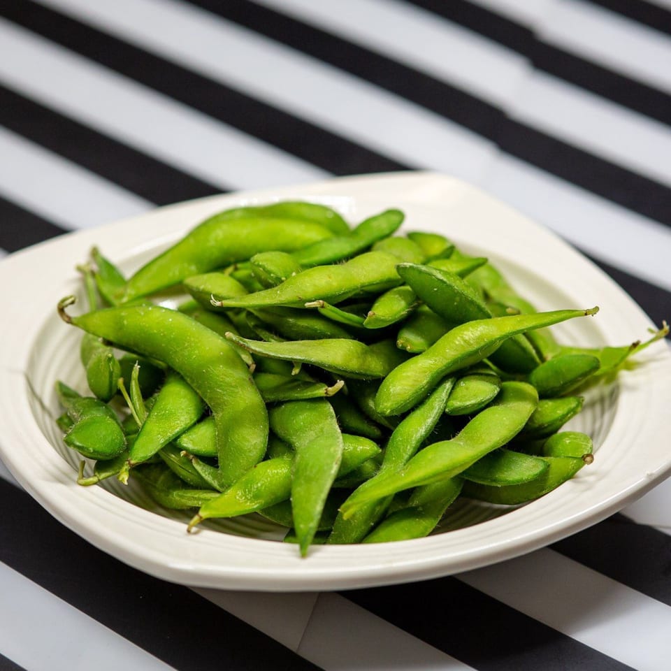 A white bowl filled with vibrant green edamame pods, lightly salted, on a striped black and white surface.