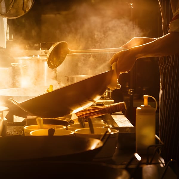 Chef stir-frying food in a wok with steam rising in a brightly lit kitchen.