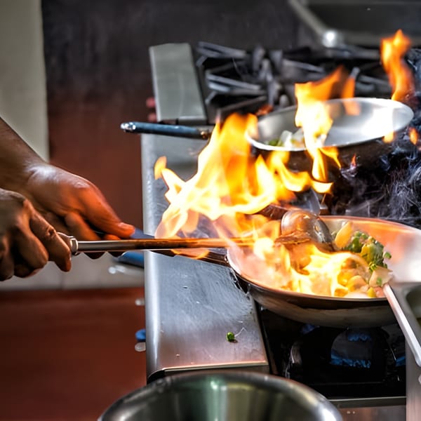 A chef's hands stir-frying food in a flaming pan on a gas stove at Nori Teriyaki Mukilteo.